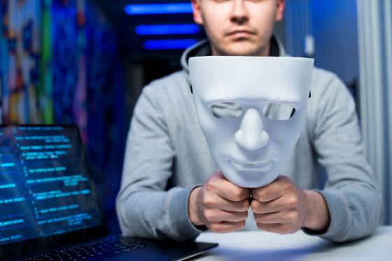 Photograph of a person holding a white, expressionless mask in front of their face, seated at a table with a laptop displaying blue code on screen, suggesting a cyber hacker. 