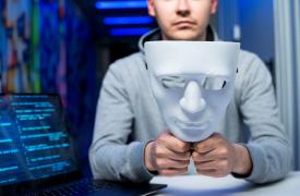 Photograph of a person holding a white, expressionless mask in front of their face, seated at a table with a laptop displaying blue code on screen, suggesting a cyber hacker. 